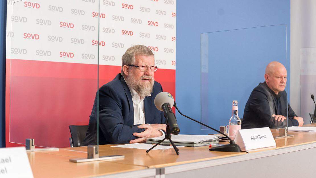 Zwei Männer sitzen mit Mikrofonen auf einem Podium. 