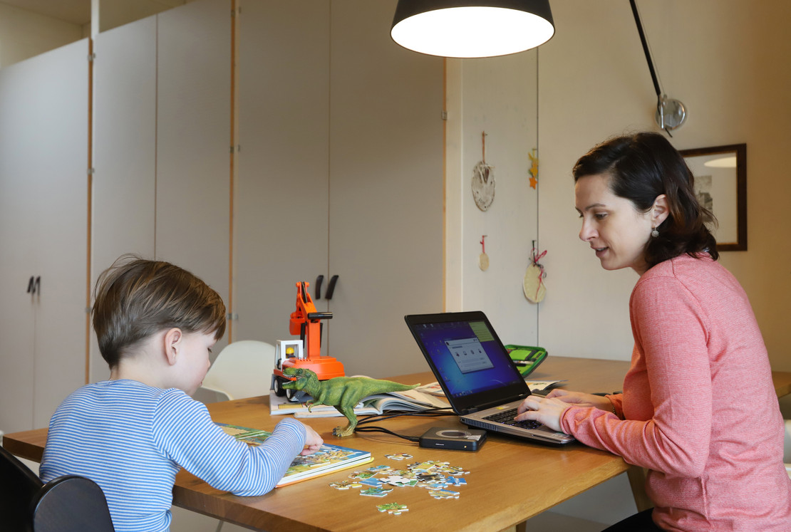 Kind spielt am Tisch, Frau arbeitet am Tisch am Laptop