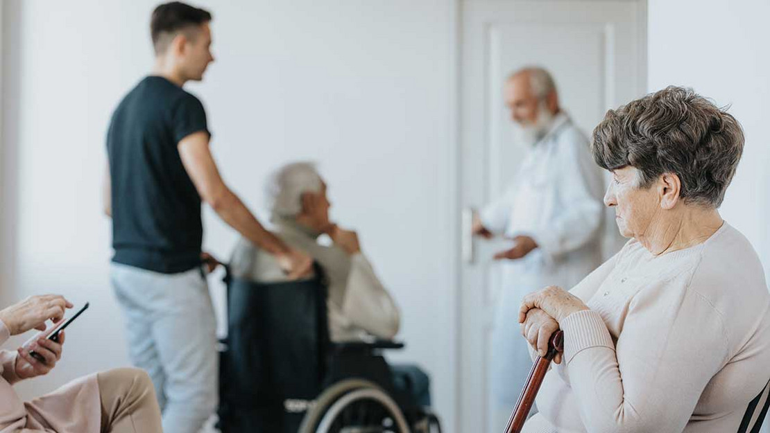 Frau sitzt im Wartezimmer, im Hintergrund wird eine Person im Rollstuhl geschoben. 