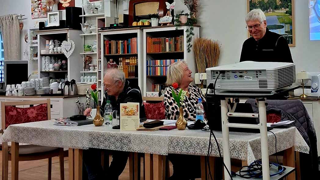 Zwei Männer und eine Frau sitzen und stehen am Tisch.