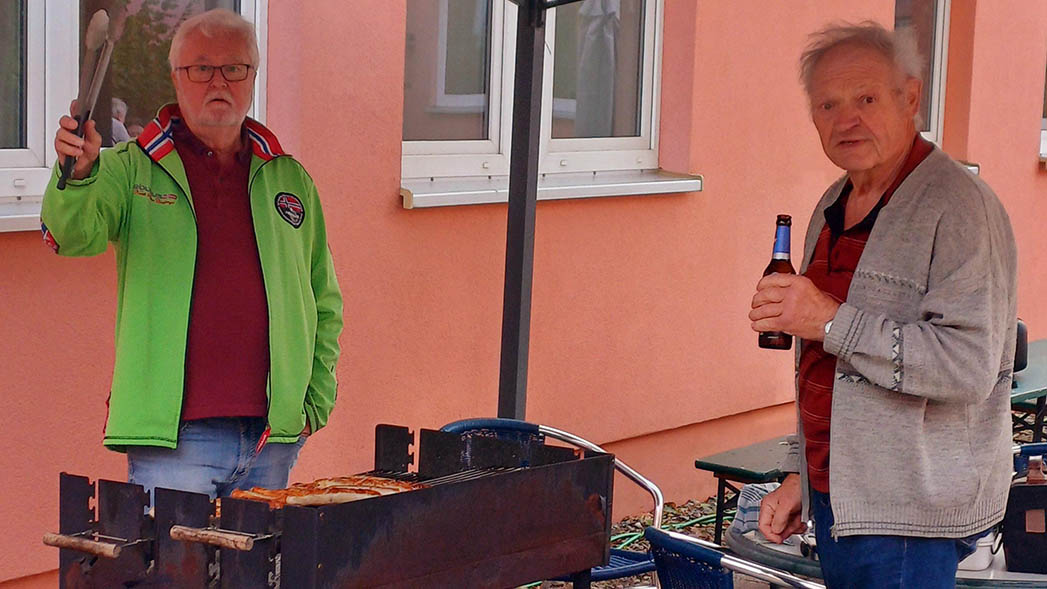 Ein Mann steht am Grill, ein anderer davor mit einer Flasche in der Hand.